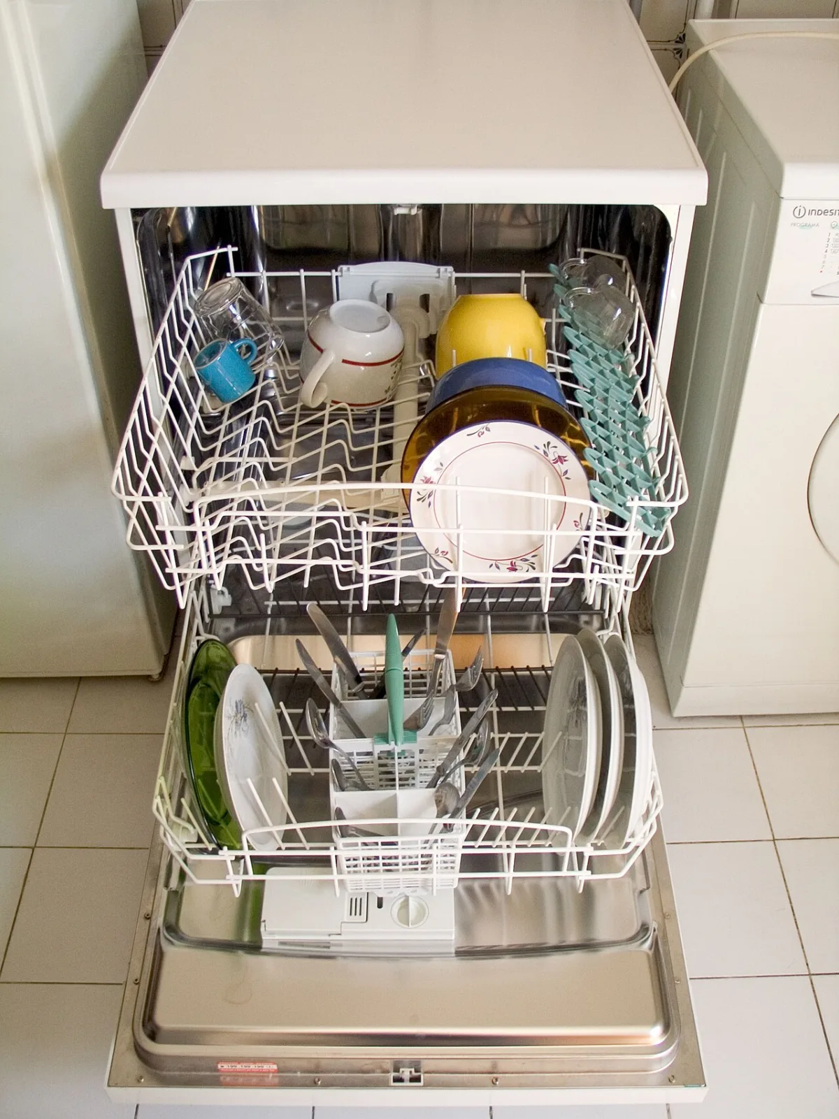 A front loading dishwasher with its door open and both racks pulled out, loaded with cups, plates and cutlery.