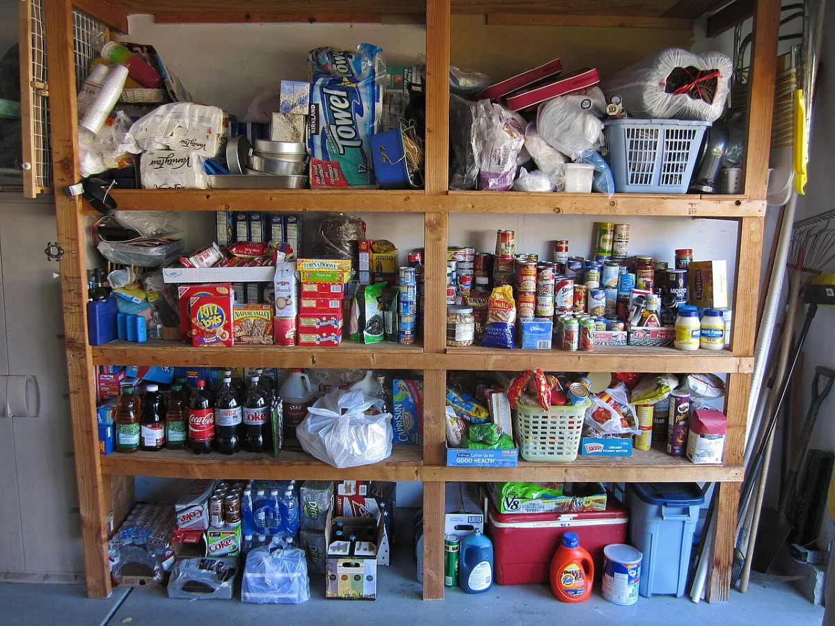 Wooden garage shelves packed with canned food, paper towels, drinks and storage bins on a concrete floor.