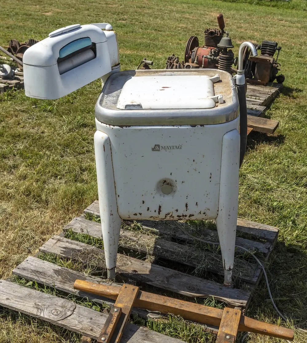 A white vintage Maytag wringer washing machine with rollers on top, sitting on a wooden pallet in the grass.