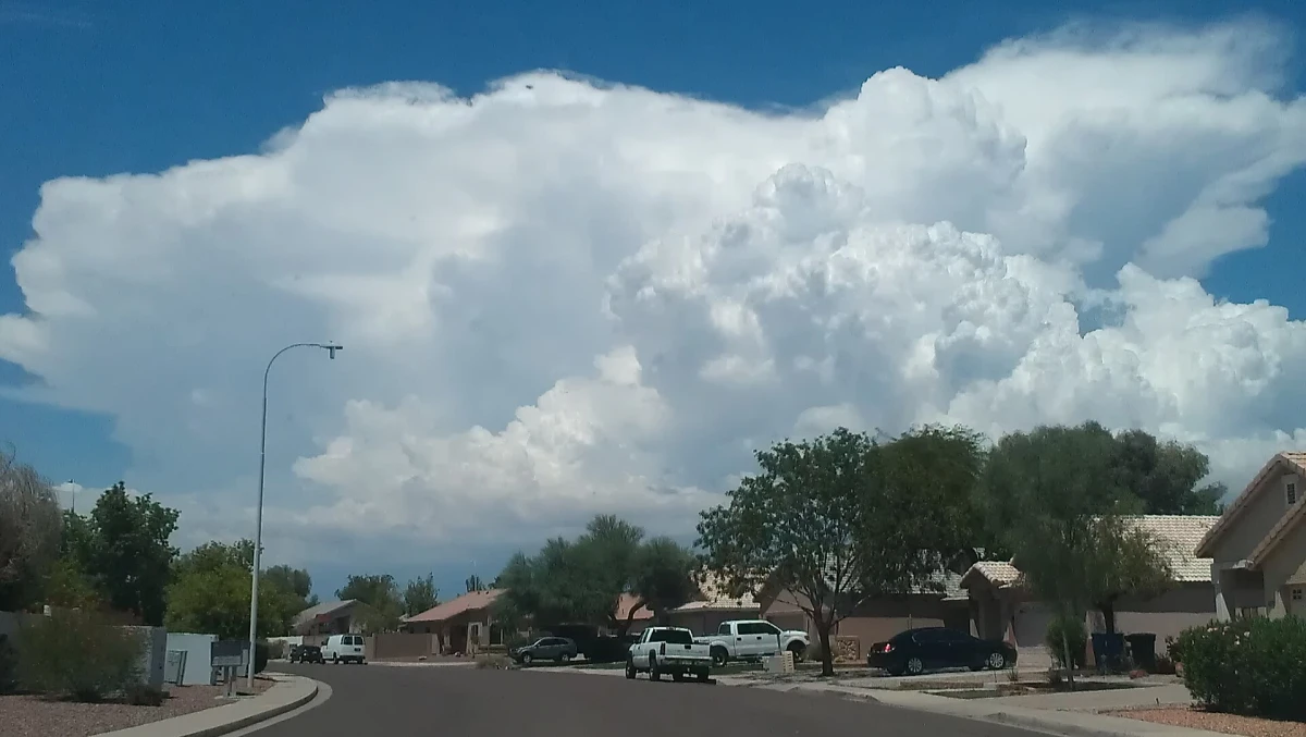 Tall white monsoon storm clouds build over a quiet Arizona street with single story homes, palo verde trees and parked trucks.