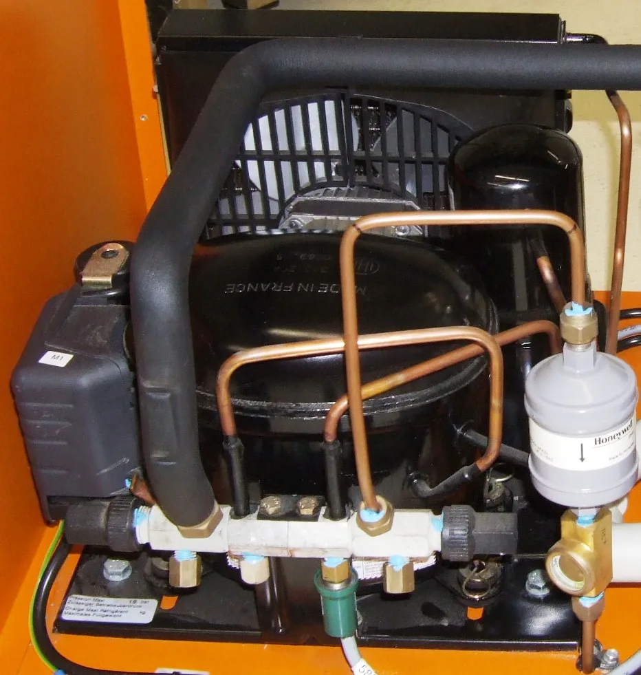 A small refrigeration condensing unit with a black compressor, copper lines and a fan behind a coil.