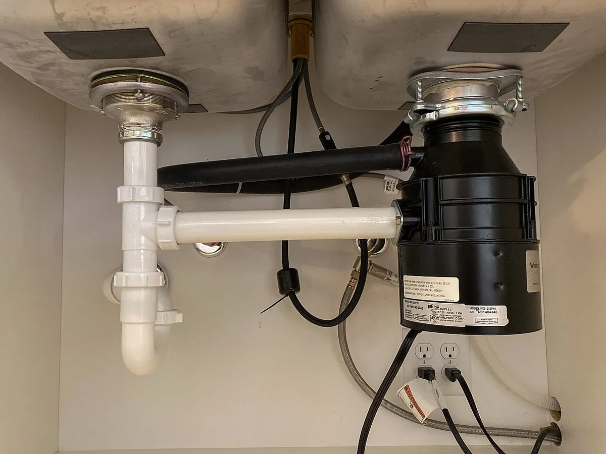 Underside of a double kitchen sink with a garbage disposal on the right, white drain pipes on the left and a dark hose entering the disposal