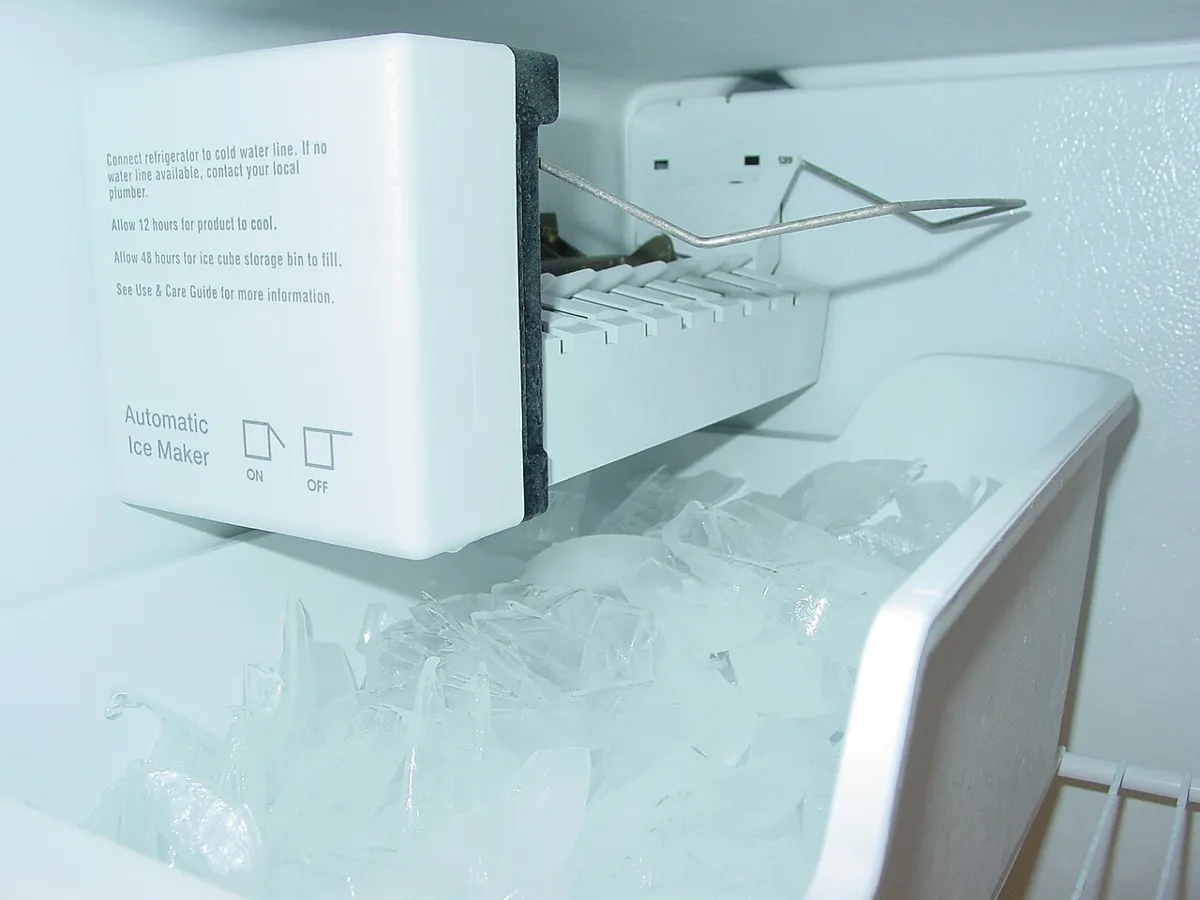 Freezer ice maker with a white cover and a wire shut off arm over a bin of ice, with the on and off positions printed on the cover