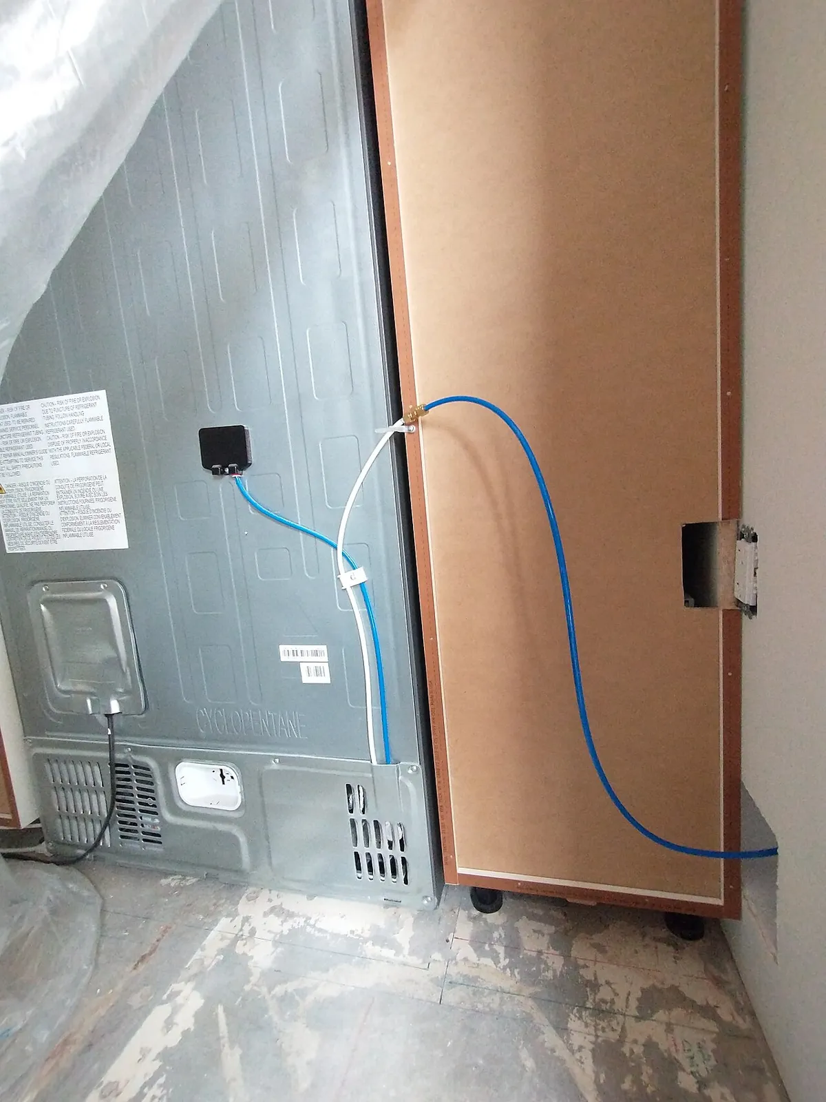 Back of a stainless refrigerator with a blue plastic water line running from the fridge to a shutoff fitting on the cabinet beside it